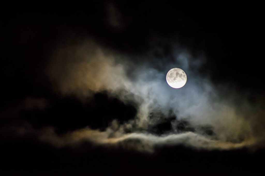 A full moon glowing brightly in a dark sky, partially surrounded by wispy clouds.