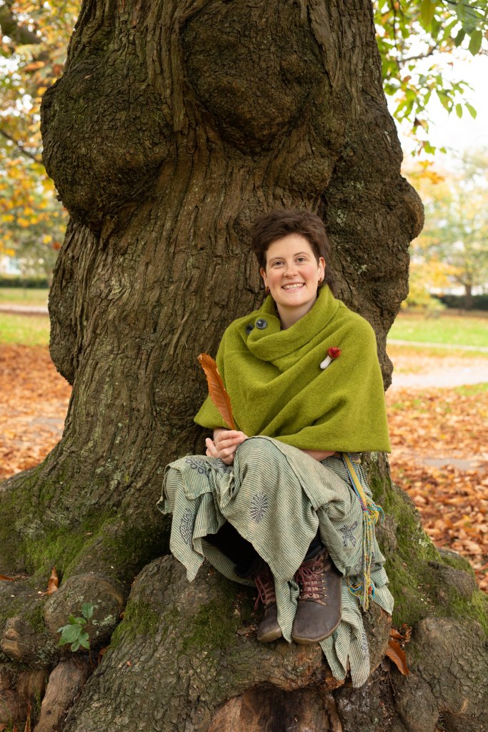 A woman sitting on the roots of a large tree, wearing a green shawl and patterned skirt, smiling warmly at the camera.