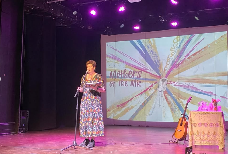 A person standing on stage holding a book while reading during an event titled 'Mothers on the Mic,' with colorful lights and a microphone graphic in the background.